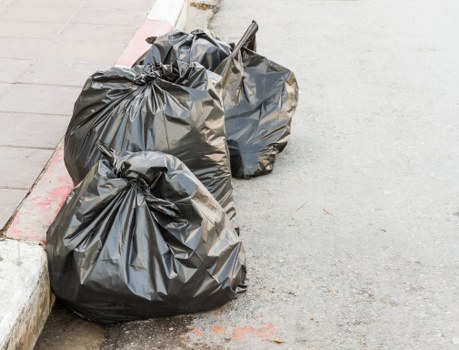 Two black plastic rubbish bags, filled with waste materials, sit on a grey concrete pavement near the edge of a curb in an urban street scene. The bags are loosely tied at the top and appear slightly overstuffed, with some bulging and creases visible on their surfaces, indicating they contain bulky rubbish. One bag is positioned closer to the foreground, while the other is slightly behind and leaning against the curb. The surrounding environment includes a section of paved sidewalk with visible cracks and a narrow strip of curb with a red painted line marking the street boundary. The scene implies a typical rubbish collection setup within a residential or commercial area, possibly in Hammersmith, London, helping to support local waste management services like those offered by Commercial Waste Hammersmith, which handles rubbish collection and disposal in the area.