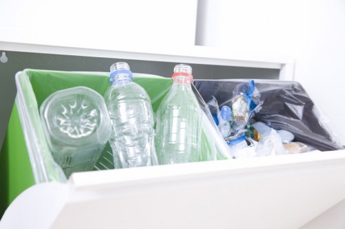 A close-up view of two transparent plastic bottles, one with a blue cap and the other with a red cap, placed inside a green plastic recycling bin, which is positioned within a white interior environment. The green bin contains additional clear plastic items such as a cup and is partly filled with other recyclable plastics, including a black plastic bag and crumpled plastic packaging, all situated on a flat surface, possibly a kitchen or utility room countertop or inside a waste collection area. The background features a white surface, likely part of a cabinet or appliance, emphasizing a clean and organized space suitable for rubbish disposal and recycling efforts. The image illustrates typical domestic waste management involving plastic bottles and small plastic containers, aligning with rubbish removal services like those offered by Commercial Waste Hammersmith in central London.