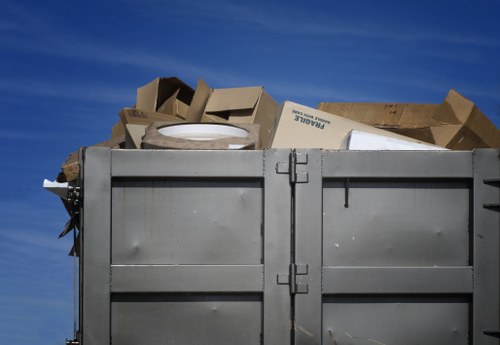 A large, grey metal skip filled with a variety of discarded cardboard boxes and packaging materials, positioned outdoors against a backdrop of a clear blue sky. The cardboard boxes are in different sizes, with some folded and others partially crushed, and display minimal printed or branding details. The containers' surface shows signs of use, with slight rust and scratches visible. The skip is situated on a flat surface, possibly a driveway or service yard, with no other objects or vehicles visible nearby. The image captures a moment of rubbish collection or disposal, typical of commercial waste management operations in Hammersmith. The focus is on the container’s contents and structure, reflecting professional rubbish removal services offered by Commercial Waste Hammersmith in the local area.