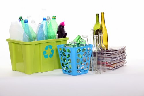 The image displays a collection of recyclable waste items organized into two plastic containers on a white surface. The larger, lime-green container on the left contains various plastic bottles made of transparent and coloured plastics, some with screw caps, and they have a smooth, glossy finish. The smaller, blue perforated basket to the right holds additional plastic bottles, primarily transparent with some green and clear ones, with visible bottle openings and labels. To the right of these containers, there are several glass bottles and jars of different shapes and sizes, including a green glass bottle, a clear glass bottle, and a tall, narrow yellow glass bottle, along with a short, rounded clear glass jar, all resting on a stack of neatly folded newspapers or pages. The overall environment appears well-lit with a neutral background, emphasizing the different textures of plastic and glass materials, supporting waste management and recycling efforts typical of rubbish removal services in the Hammersmith area.