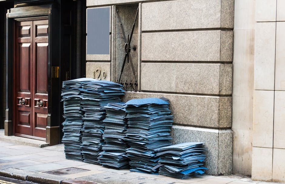 A large pile of disorganized blue-and-white paper documents stacked against a stone wall outside a building entrance, with some stacks leaning slightly. The documents appear to be standard letter-size