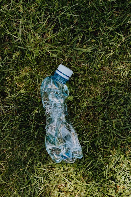 A crushed clear plastic water bottle with a white screw cap lies on a grassy surface, partially flattened with visible creases and folds in the plastic. The bottle appears to be intended for disposal 